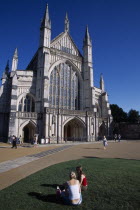 Winchester Cathedral exterior seen from the gardens with two girls sat on the grass in the foreground.