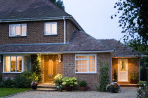 Semi detached house brick built and garden with gravel driveway at dusk with lights on in windows and above external doors