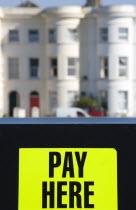 Yellow car park sign with black writing saying Pay Here with terraced seafront houses in the background