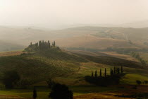 ITALY, Tuscany, San Quirico D'Orcia, The Belvedere Building on a hilltop surrounded by cypress trees sitting above olive groves and wheat fields in Val D'Orcia valley at sunrise on a misty morning.