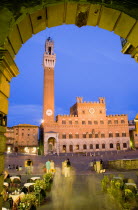 ITALY, Tuscany, Siena, Early evening in the Piazza del Campo seen through an archway with people at restaurant tables and walking in the square. The illuminated Torre del Mangia, the second tallest be...