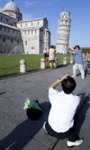 ITALY, Tuscany, Pisa, Asian male tourist taking a photograph of another Asian man pretending to hold up the Leaning Tower of Pisa in The Campo dei Miracoli or Field of Miracles.