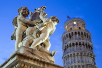 ITALY, Tuscany, Pisa, An illuminated statue in The Piazza del Duomo of cherubs holding a shield bearing the Cross of Pisa with the Leaning Tower beyond.