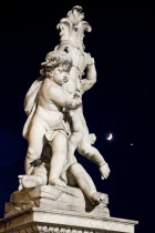 ITALY, Tuscany, Pisa, An illuminated statue in The Piazza del Duomo of cherubs holding a shield bearing the Cross of Pisa with a half crescent moon shining in the night sky beyond.