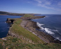 View east from the Worm s Head towards the mainland.  Sloping ridge of cliff extending from mainland with waves breaking against side.