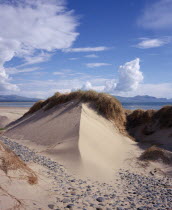 Wind shaped sand dune topped with thatch of grasses and with smooth grey pebbles scattered across the sand in the foreground.