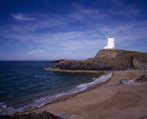 Curving  empty beach overlooked by old lighthouse standing on rocks extending out to sea.  Blue sky and windswept clouds above.