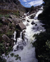Ogwen Falls and Nant Ffrancon Pass.   White foaming water tumbling over rocks with arched stone bridge in background.