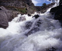 Ogwen Falls and Nant Ffrancon Pass.   White foaming water tumbling over rocks with arched stone bridge in background.