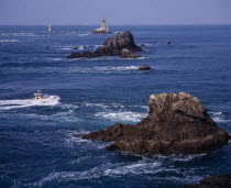 Pointe du Raz.  Jagged outcrops of offshore rock protruding from sea with small fishing boat facing oncoming tide  light house and Ile de Sein skyline.