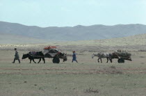 Nomad family leading yak and horse drawn carts laden with their belongings through open landscape.