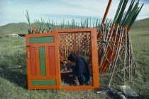 Man painting interior framework of yurt sitting within circular lattice structure framed by newly painted open doorway.