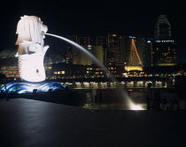 The Merlion statue at the Merlion Park river entrance at night with the esplanade concert hall  Oriental Hotel and other city buildings illuminated behind.