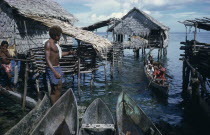 Thatched stilt houses on artificial island with man and child on wooden bridge and approaching canoe.  Part view of wooden canoes in immediate foreground.