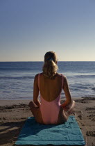 Young woman on sandy beach sitting in a meditation position on a green towel looking out towards the sea