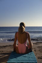 Young woman sitting on a sandy beach wearing a pink swimsuit with back to camera looking towards the sea