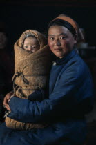Young mother in fleece-lined silk tunic holding baby wrapped in traditional swaddling against bitter winter cold.East Asia Asian Babies Classic Classical Historical Kids Mongol Uls Mongolian Mum Olde...