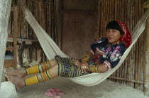 Young Kuna mother rests in hammock outside home  wearing brightly coloured traditional layered applique mola panel    traditional bead design arm and leg bands and gold nose ring.Cuna Caribbean Ameri...