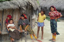 Kuna Indian family with women wearing brightly coloured tradtional mola panel blouses or dulemolas outside their thatched home  their legs bound with traditional design strings of coloured glass beads...