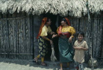 Kuna Indian women wearing brightly coloured traditional mola panel blouses or dulemolas joking outside their home with young boy standing at side.Cuna Caribbean American Central Central America Color...