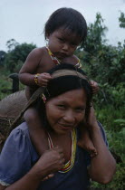 Makuna mother carrying her baby son on her shoulders and basket on her back supported by head strap.  Both  wearing necklaces of white red and yellow glass beads.Tukano  Makuna Indian North Western A...