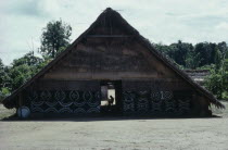 Makuna communal home or maloca with white and yellow clay and  charcoal painted design on exterior facade.  Man basket-making silhouetted in rear womens entrance.Tukano  Makuna Indian North Western Am...