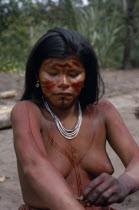 Young Barasana mother using cumare fibre bound twigs to apply red ochote fruit  line pattern to upper body and arms.  Hands already coloured dark purple with we leaf dye to wrists.  Wearing white yell...