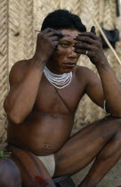 Barasana man looking in mirror to apply red ochote facial paint   thick dark purple we leaf juice vertical stripes painted on legs  wearing multi stranded white glass bead necklace. Tukano sedentary...