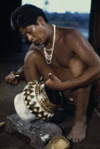 Barasana tribesman painting white yellow clay & black charcoal design onto yaje pot used to hold hallucinogenic yaje juice from vine species Banisteriopsis drunk during shamanic and religious ceremoni...