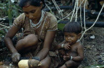 Barasana woman peeling manioc roots with small child sitting on ground beside her with painted face and wearing white glass bead necklace and bracelets.Tukano sedentary Indian tribe North Western Ama...