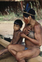 Maku hunter  face painted with red ochote marks and wearing  macaw feather crown in preparation for dance  sits beside young boy.indigenous tribe indian nomadic American Colombian Columbia Hispanic I...