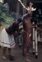Maku woman painting the body of man wearing feather crown with red karajuru/ochote plant dye.indigenous tribe indian nomadic American Colombian Columbia Female Women Girl Lady Hispanic Indegent Latin...
