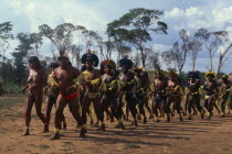 Panar men and women performing traditional dance around central area of relocated village in Xingu National Park.  Men wearing head-dresses or crowns of feathers and face and body paint.Formally kno...