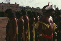 Panara men wearing crowns or head-dresses of feathers beside women painted with red karajuru and wearing colourful strands of beads and with long strips of plant fibres tied around upper arms during d...