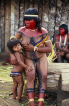 Panara woman breastfeeding daughter before dance  with face and neck painted with red karajuru and wearing multi-strand of beads across upper body and long strips of plant fibres tied around upper arm...