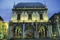 Piazza della Loggia.  Facade of Loggia or town hall with arched entrance and colonnades  balcony and statues illuminated at night.  With poster advertising Monet exhibition