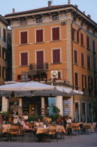 People sitting beneath white awning at outside tables of cafe in piazza of town beside Lake Garda.   Facade of building behind painted orange with red window shutters.