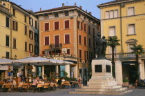Italy, Lombardy, Salo, people sitting at outside cafe tables in piazza of town beside Lake Garda. Facades of buildings  bank and hotel painted pale yellow and orange with window shutters.