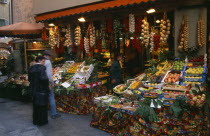 Member of staff and customers outside fruit and vegetable stall with pavement display and strings of onions and garlic hanging from rack above doorway.