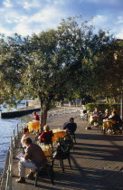 Lake Maggiore.  People enjoying lakeside views sitting at cafe tables beneath trees on the esplanade.  Late afternoon sun and long shadows.