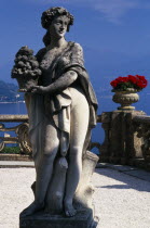 Villa Balbianello.  Statue of woman holding basket of fruit with red geraniums in stone vessels on stone balustrade behind and view over lake.