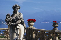 Villa Balbianello.  Statue of woman holding basket of fruit with red geraniums in stone vessels on stone balustrade behind and view over lake.