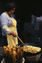 Woman using long pair of chopsticks to turn frying food in large  shallow dish in front of her.