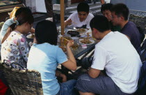 Wangfujing shopping street.  Group of young people using chopsticks to eat meal  and drinking at outside table.