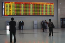 Interior of railway terminal with electronic information board with customers looking at smaller screens beside ticket machines below.