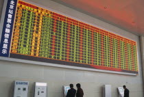 Interior of railway terminal with electronic information board with customers looking at smaller screens beside ticket machines below.