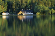 Austria, Oberosterreich, Mondsee, view across lake towards boat house and other building with forest backdrop reflected in flat surface of water.