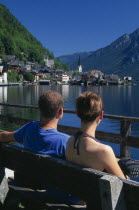 Young couple sitting on wooden bench looking out across Hallstattersee Lake towards village church  houses and boathouses with mountain backdrop.