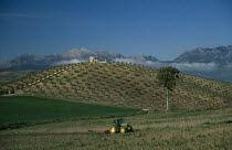 A tractor ploughing a terrace overlooked by a stone building on a hill and mountains with mist in the distance
