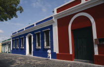 Red  blue and white painted house frontages near Plaza Bolivar with a person walking past